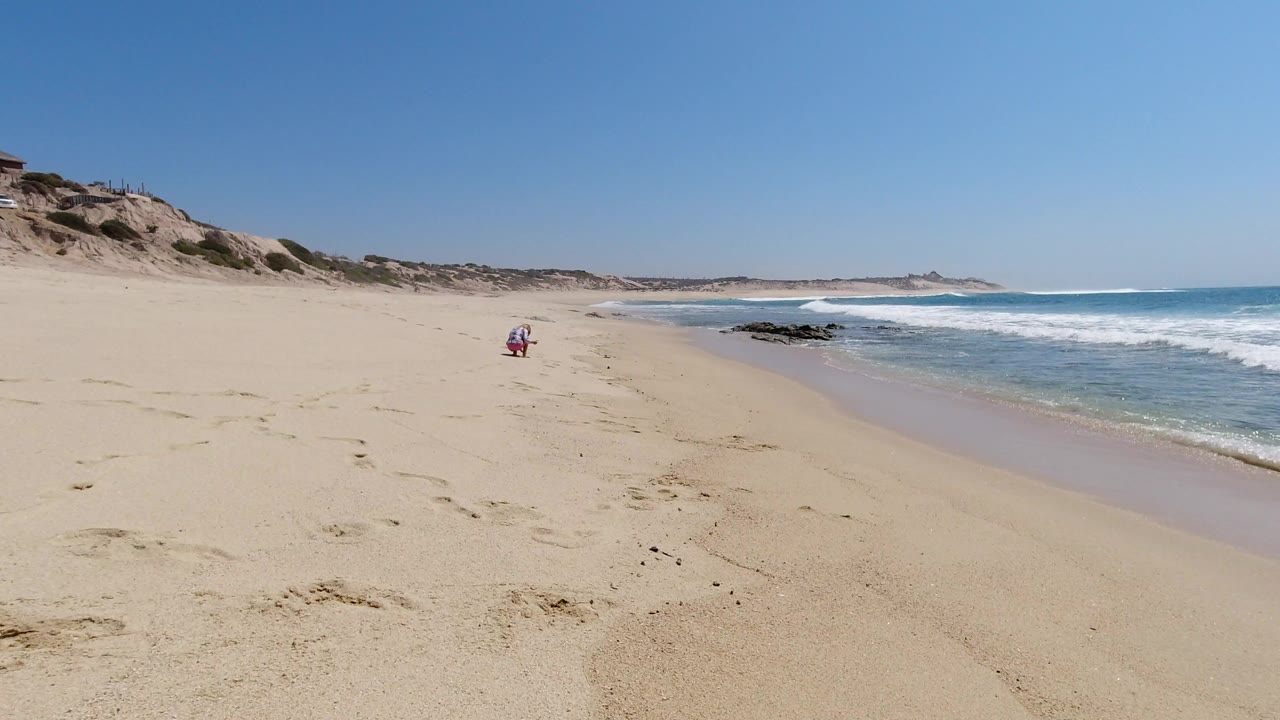 Los Cabos shoreline view with the waterline slightly farther into frame, dark rocks near the surf, dune-backed sand, and no visible people.