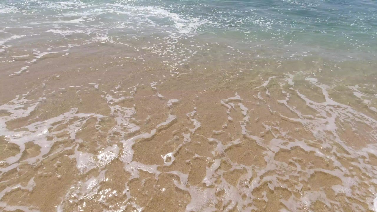 Small foamy waves cover wet tan sand in Los Cabos, Mexico, with blue-green water near the top of frame and no visible people.