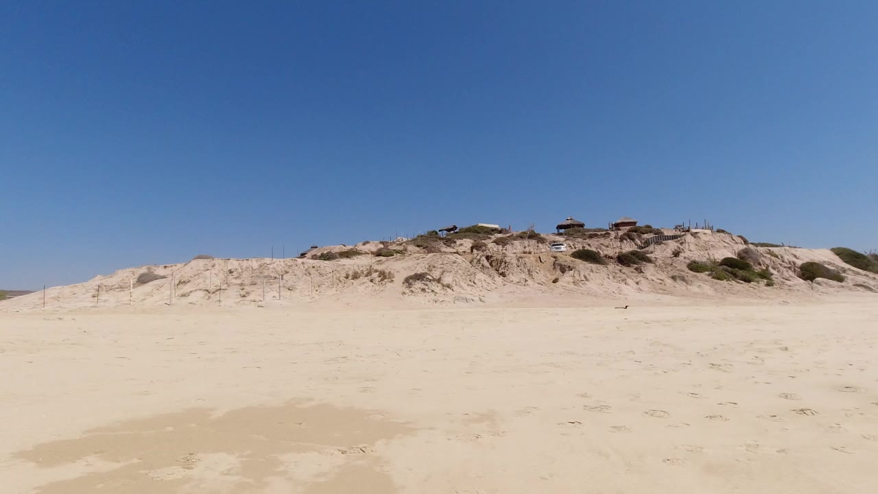 Wide Los Cabos beach view with pale sand in the foreground, low dunes and small structures along the horizon, and no visible people at the start of the clip.