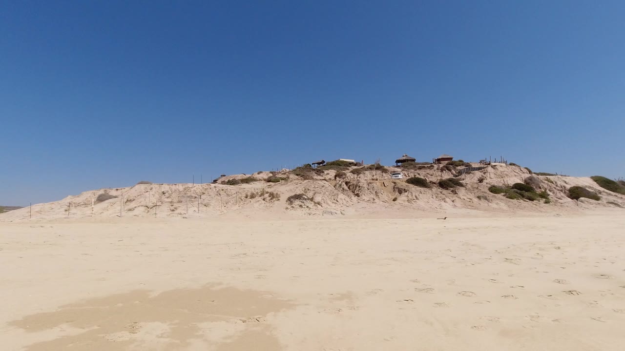 Los Cabos beach frame with empty sunlit sand, a low dune ridge across the background, small distant structures, and no visible people.