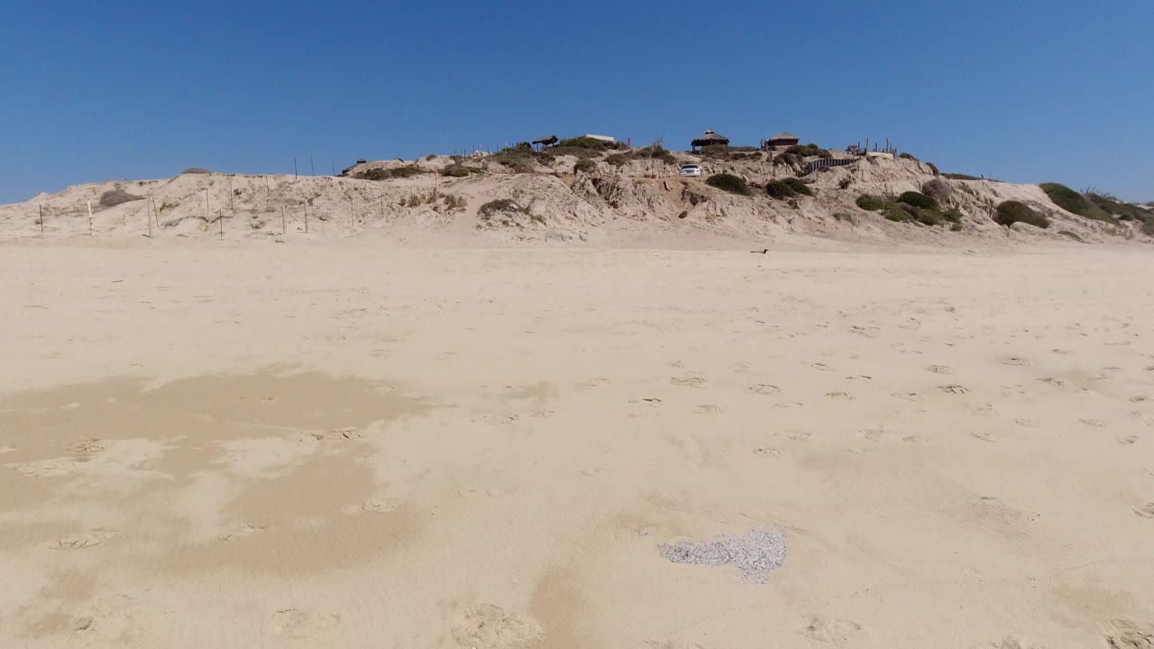Opening frame of an empty sandy beach in Los Cabos, Mexico, with pale dunes, sparse scrub, ridge-top structures, and no visible people.