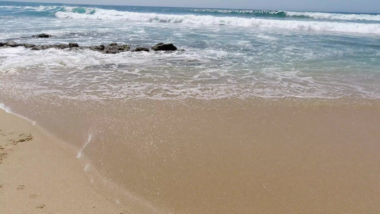 Los Cabos shoreline with white waves breaking behind dark rocks, wet tan sand in the foreground, blue horizon, and no visible people.