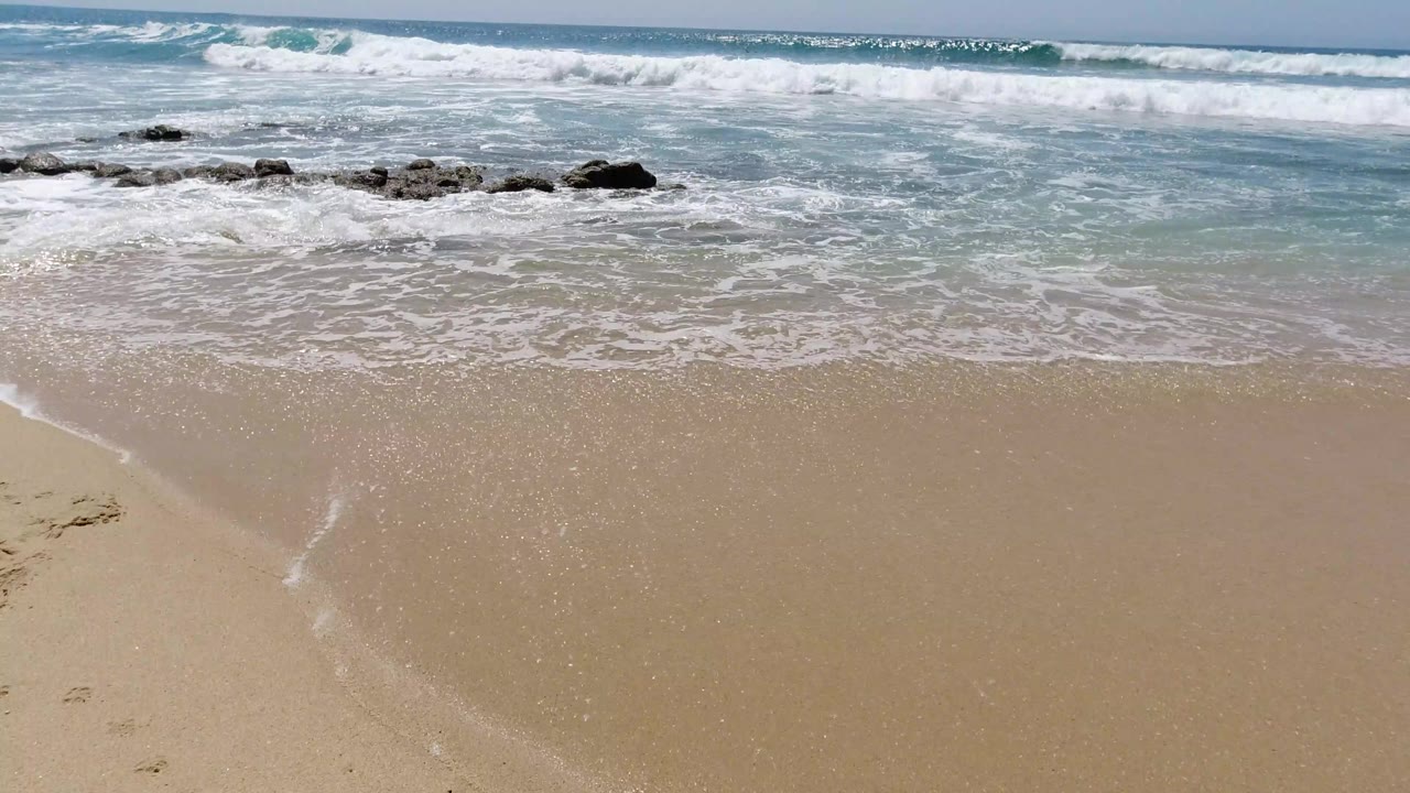 Los Cabos beach view with a thin sheet of foam reaching farther onto wet sand while waves break beyond the low rocks, no visible people.