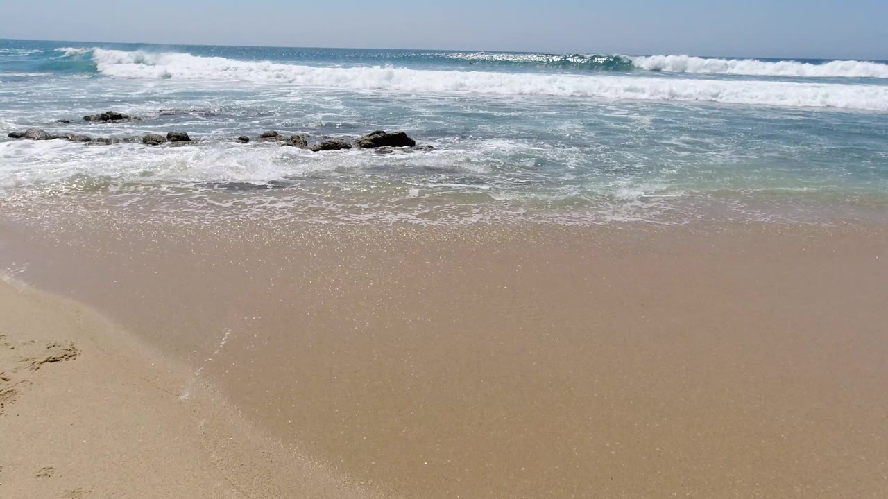 Wide shoreline view with surf, rocks, and smooth sand.