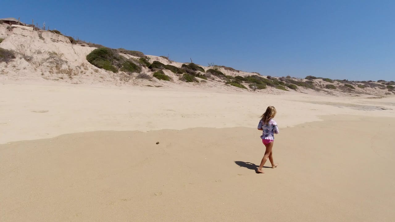 A girl walks across open sand in Los Cabos, Mexico, with pale dunes and sparse greenery behind her and no ocean visible yet in this opening frame.