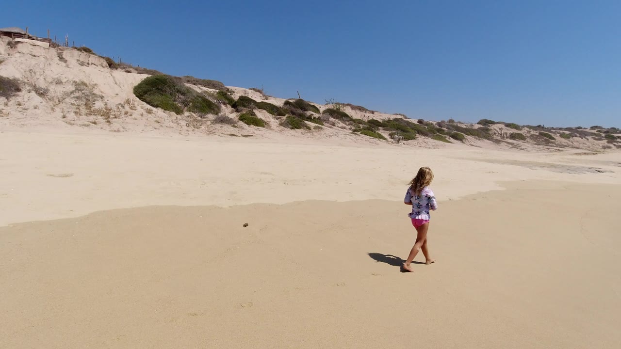 The girl continues along the wide beach in Los Cabos, Mexico, with low dune cliffs across the background and hard sunlight casting a dark shadow on the sand.