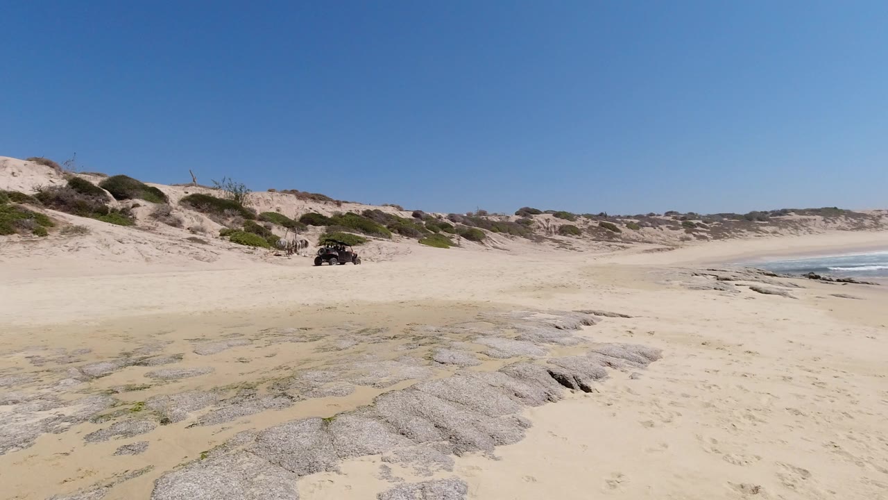 Wide Los Cabos beach view with pale sand, exposed rock shelves, low dunes, ocean surf at right, a small dark vehicle in the distance, and no visible people.