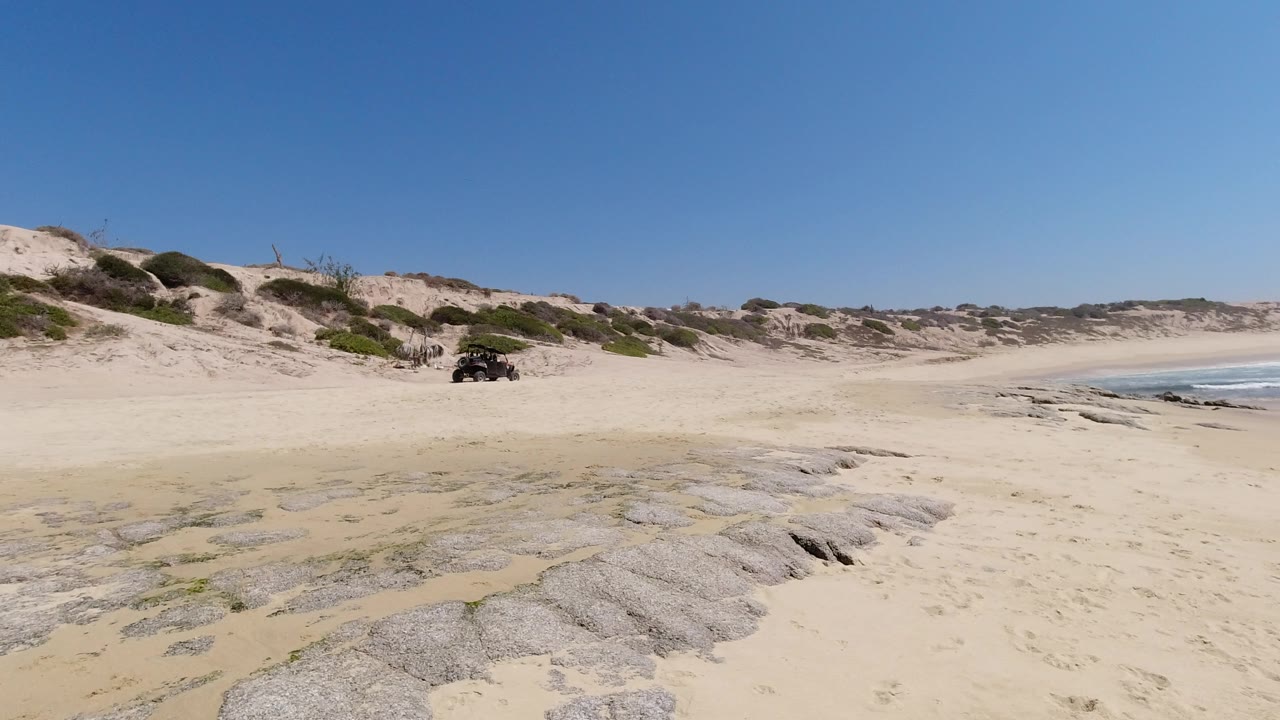 Wide Los Cabos shoreline frame with rock shelves in the foreground, a dark off-road-style vehicle near the dunes, blue sky above, and no visible people.