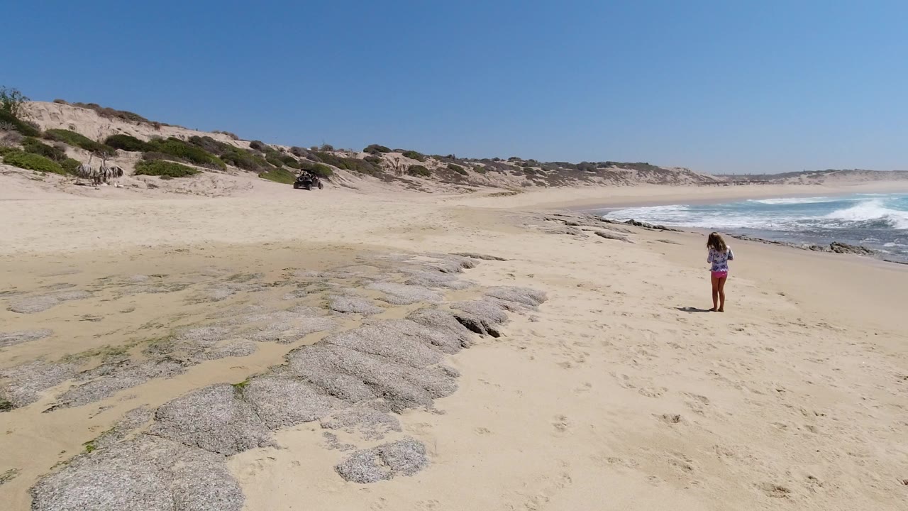 Wide beach view with a woman standing near the surf and dunes behind her.