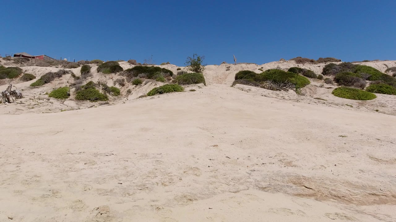 Wide sandy terrain below scrub-covered low hills and blue sky.