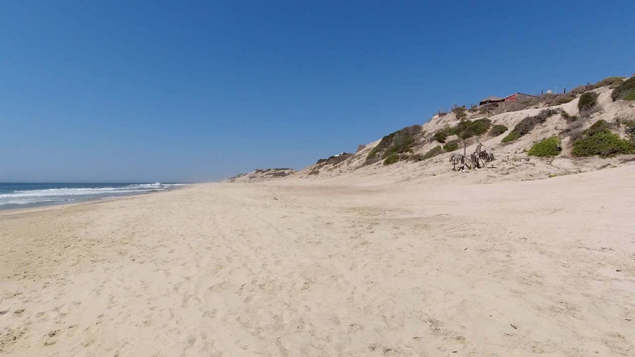Los Cabos beach frame showing open sand, low waves along the left edge, dune slopes with green brush to the right, and no visible people.