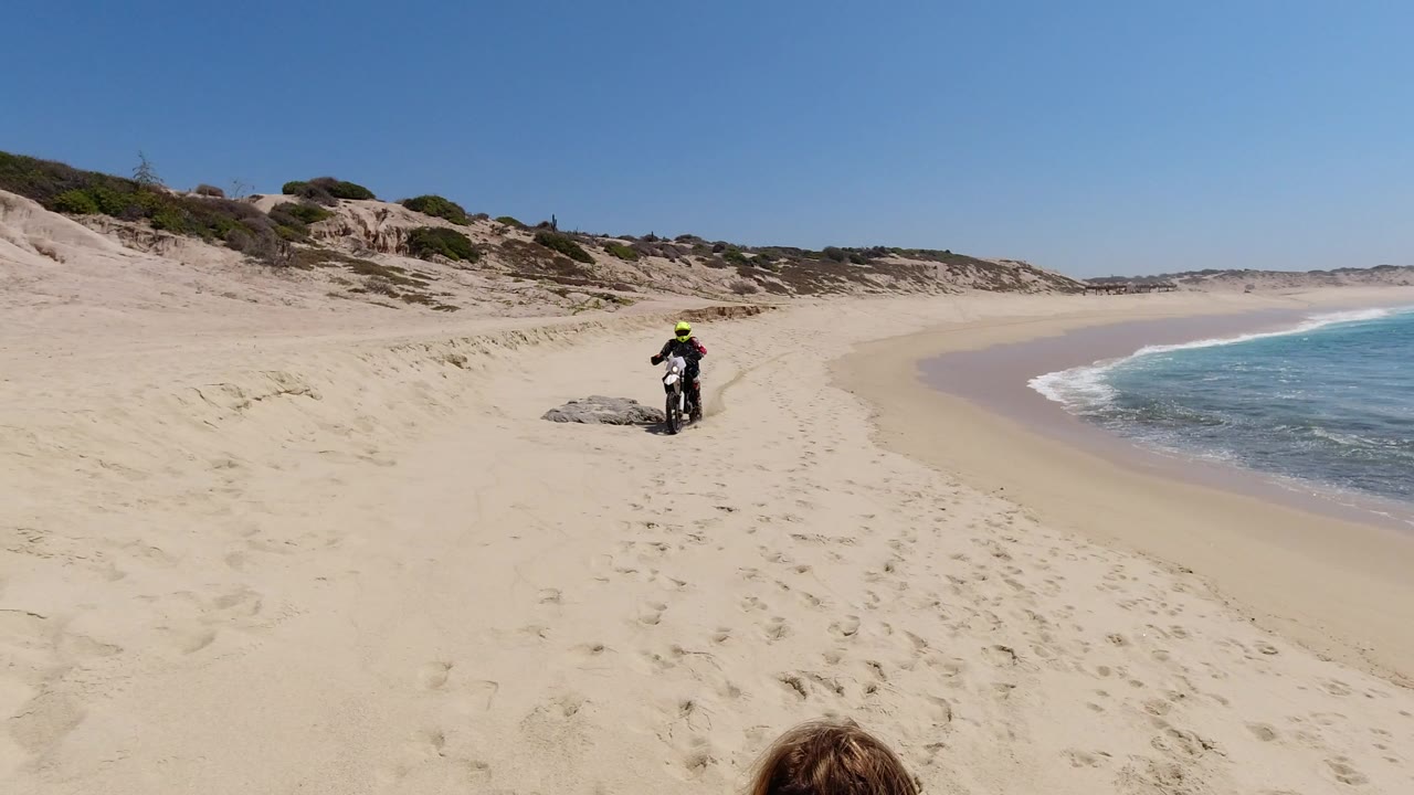 The motocross rider continues across pale sand in Los Cabos, with the shoreline curving along the right edge and dune scrub rising in the background.