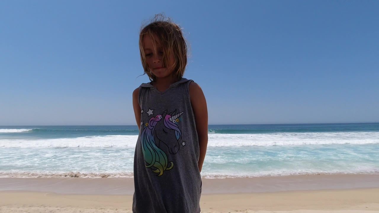 Young blond girl standing on a sunny beach with ocean waves behind her.