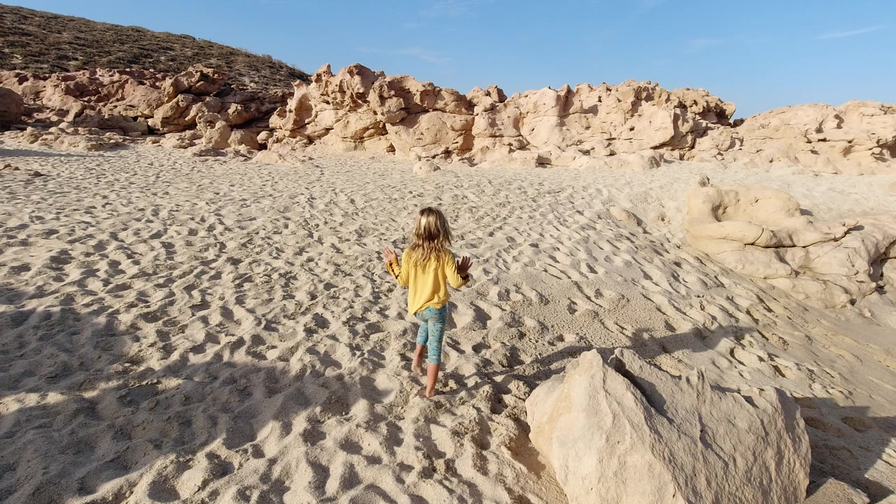 Young girl walking barefoot across rippled sand toward pale rocks.