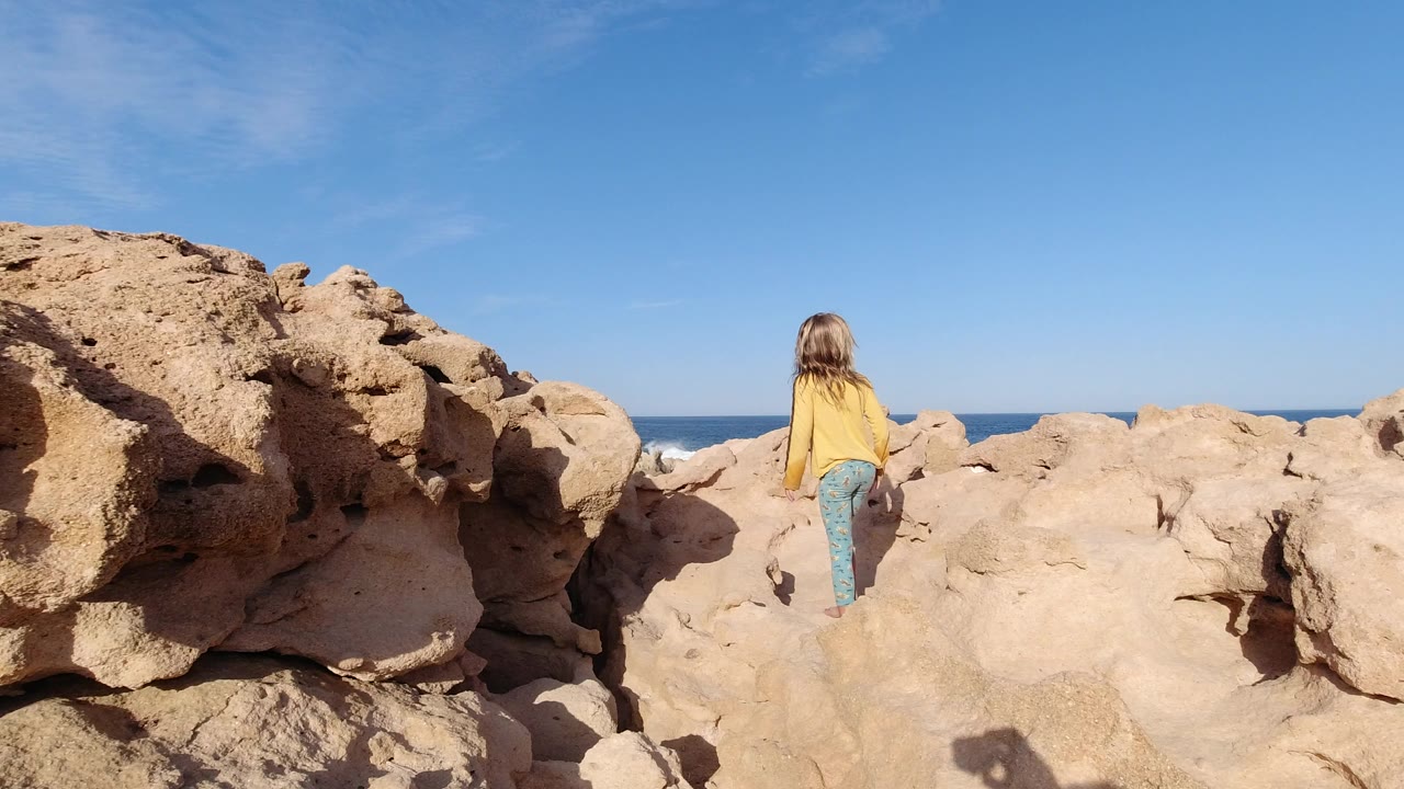 Young girl walking away across tan rocks beside the ocean.