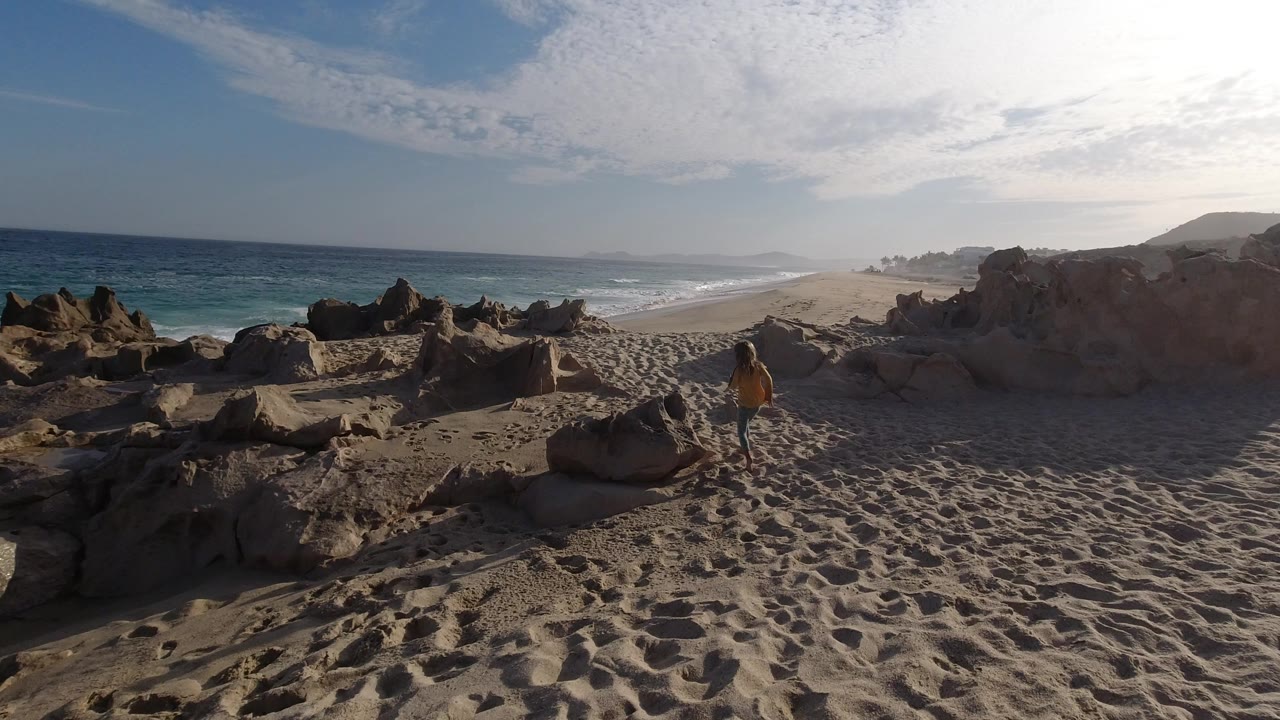 Wide Los Cabos beach frame with pale sand, dark rocks in the foreground, blue surf at left, and a young girl in yellow standing near the center-right path.