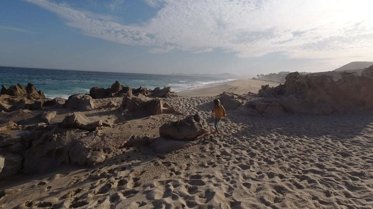 Los Cabos shoreline view with the young girl stepping between sandy rocks, ocean waves rolling in from the left, and hazy beach stretching into the distance.