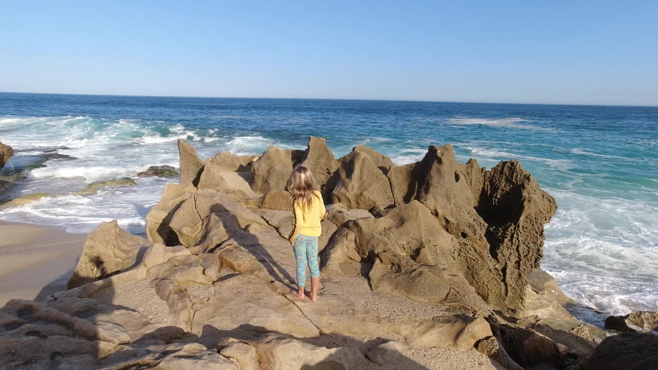 Young girl standing barefoot on tan rocks above ocean surf.