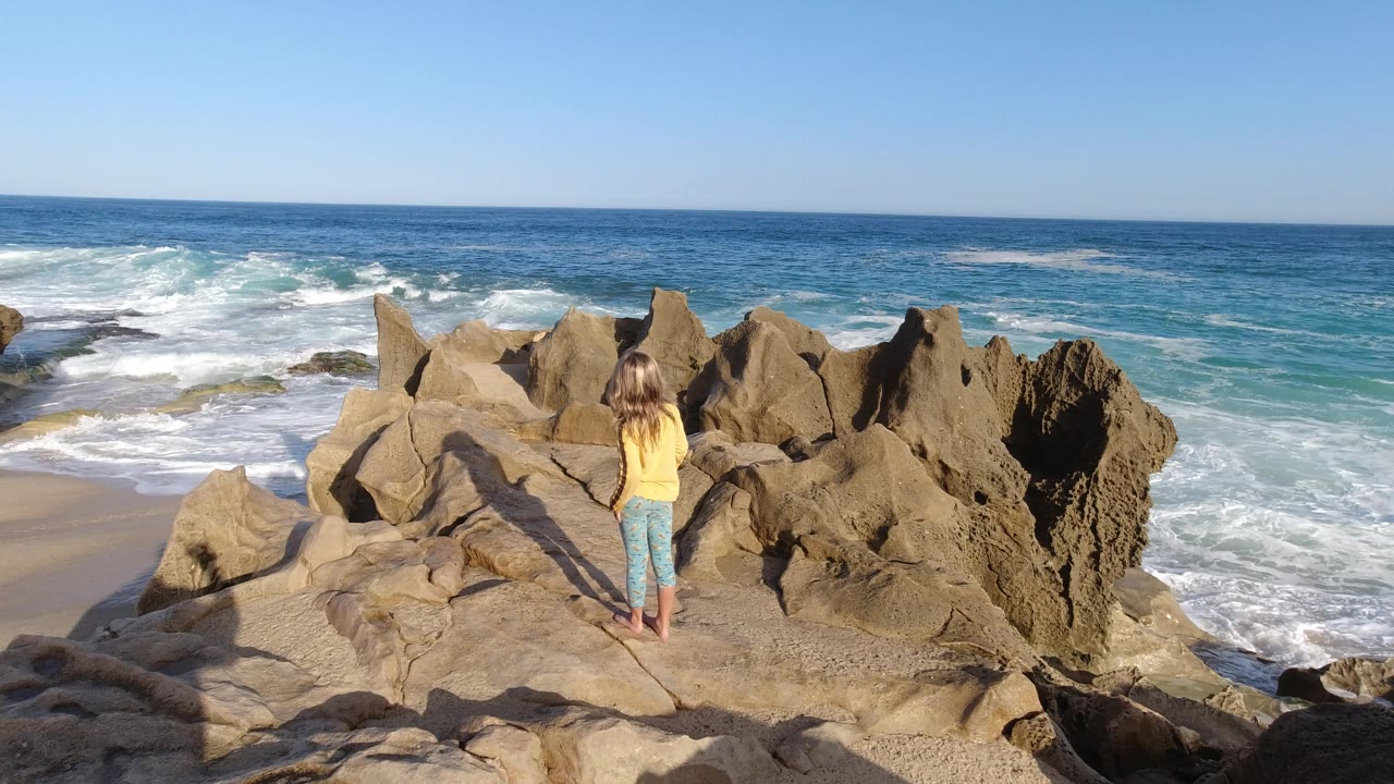 At 0:01 in Los Cabos, Mexico, the girl remains centered on the rocks while foamy water spreads along the shoreline behind the jagged stone formations.