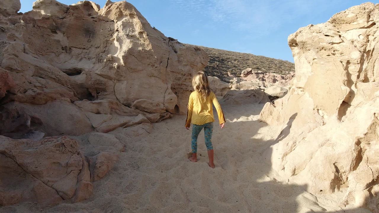 Girl in a yellow coat standing on sandy ground between pale rock formations.