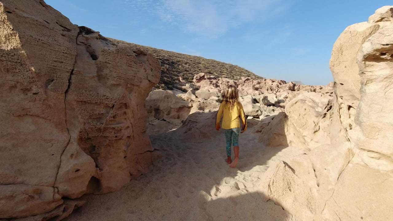 A long-haired child walks away through a narrow sandy passage between pale rocks in Los Cabos, Mexico, with bright blue sky above.