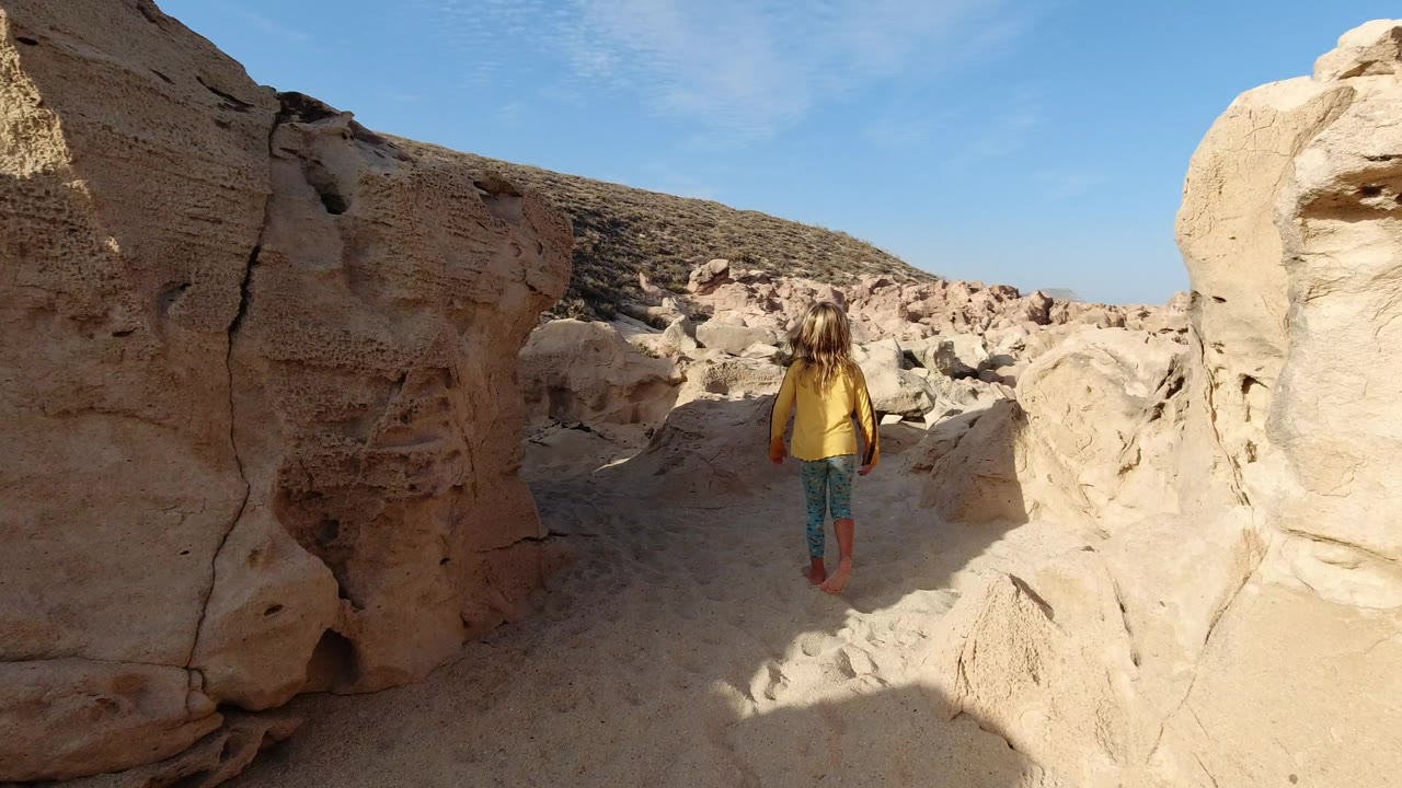The child continues away from the camera through pale eroded rocks in Los Cabos, Mexico, with a scrubby hillside visible ahead.