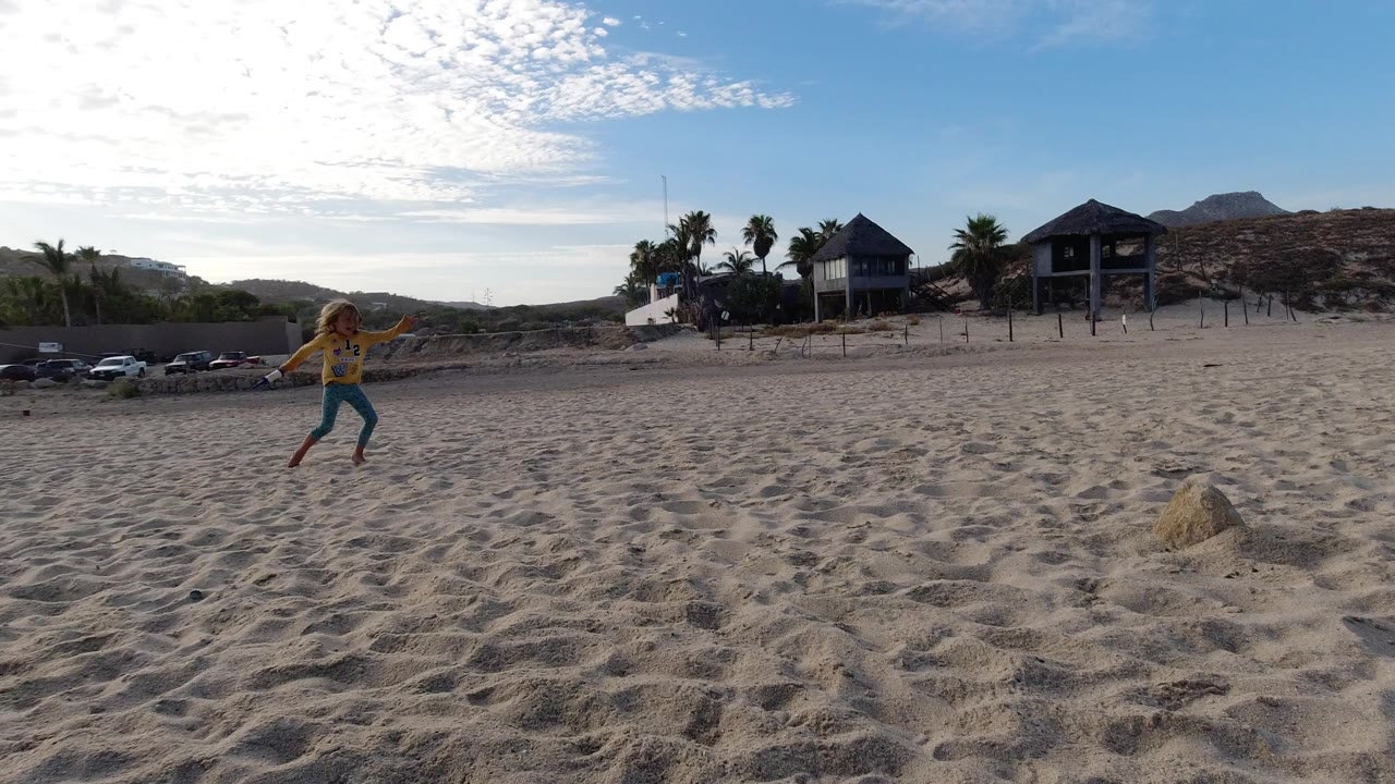Child runs across sandy beach with beach houses and hills behind.