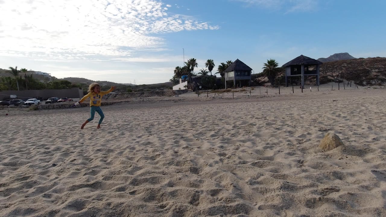 The child moves closer to center frame on the Los Cabos beach, with textured sand in the foreground and houses, palms, cars, and hills across the horizon.