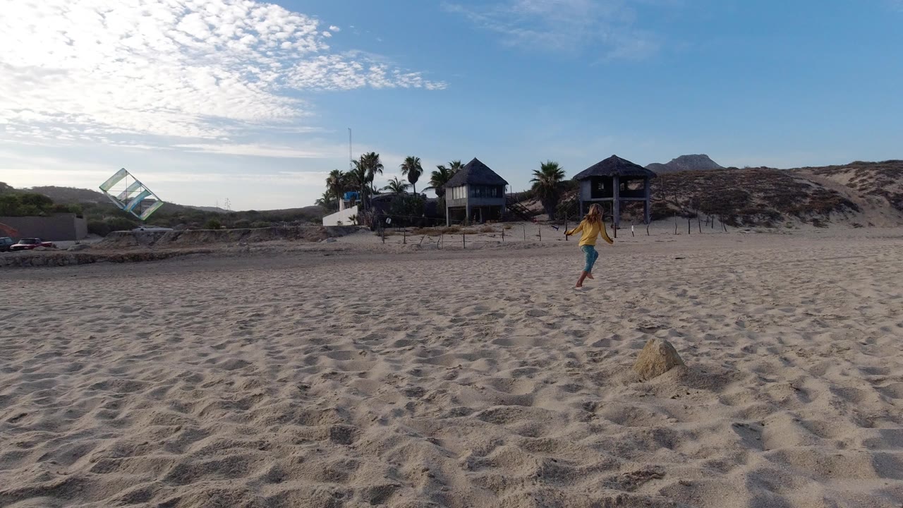 Child runs on a sandy beach with a colorful kite at left and beach houses behind dunes.