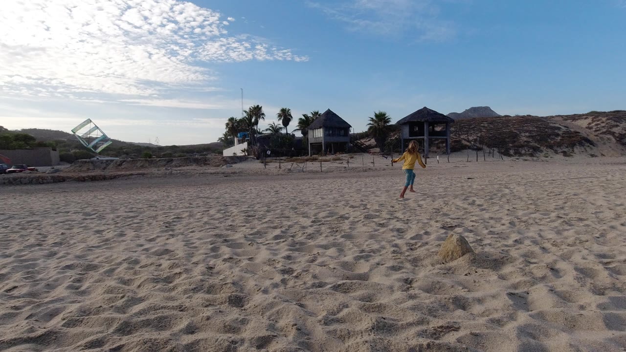 At 0:01 in Los Cabos, Mexico, the child is farther across the sand, with the colorful kite low on the left and a row of palms and raised beach houses behind the dune line.