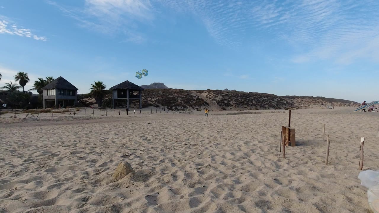 Wide Los Cabos beach view at the start of the clip, with rippled sand, wooden stakes, two thatched structures near dunes, distant people, and kites in a clear blue sky.