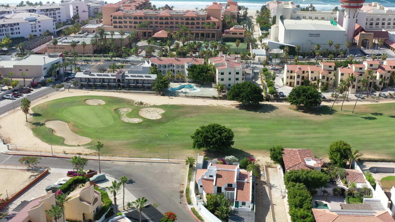Opening aerial frame over a Los Cabos golf course with green fairway, sand bunkers, nearby homes, resort buildings, palm trees, and no visible people.