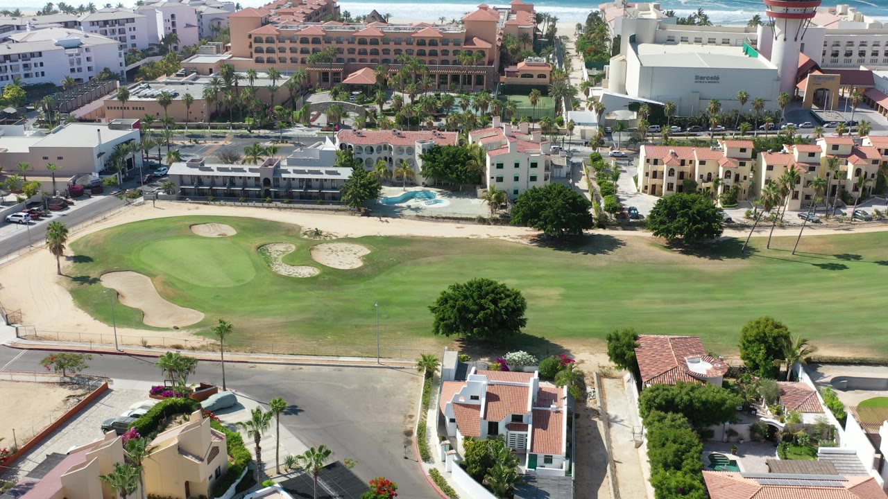 Aerial view in Los Cabos showing a bright green course, curved sand traps, roadside homes, larger hotel buildings, distant surf, and no visible people.
