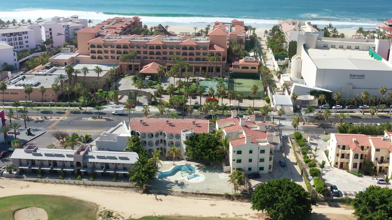 Early frame of Los Cabos, Mexico from above, showing terracotta-roofed resort buildings, palm-lined streets, pale sand, turquoise ocean, and no visible people.