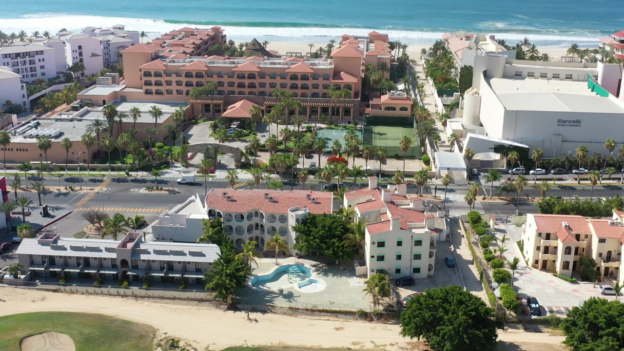 Los Cabos aerial frame with the white hotel block shifted nearer the right side, resort roofs centered, surf behind the beach, and no visible people.