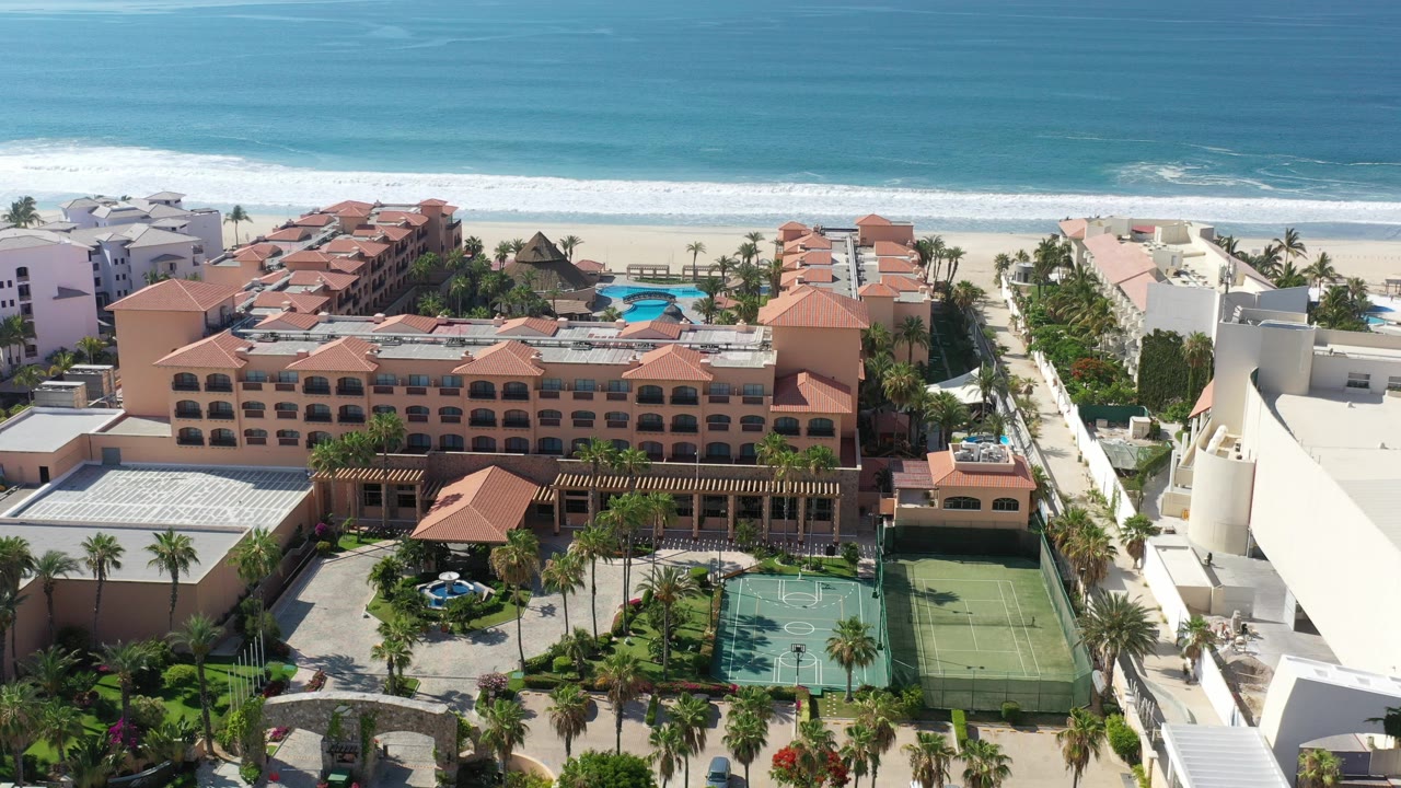 Los Cabos aerial frame showing terracotta resort roofs, palm landscaping, tennis courts, pale beach, and blue ocean surf with no visible people.