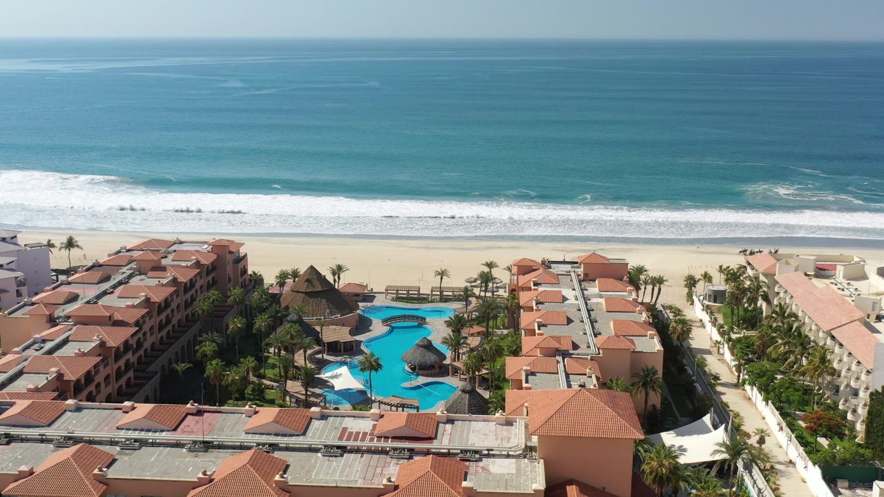 Opening aerial frame over Los Cabos, Mexico showing red tile resort roofs, blue pools, a pale beach, white surf, turquoise water, and no visible people.