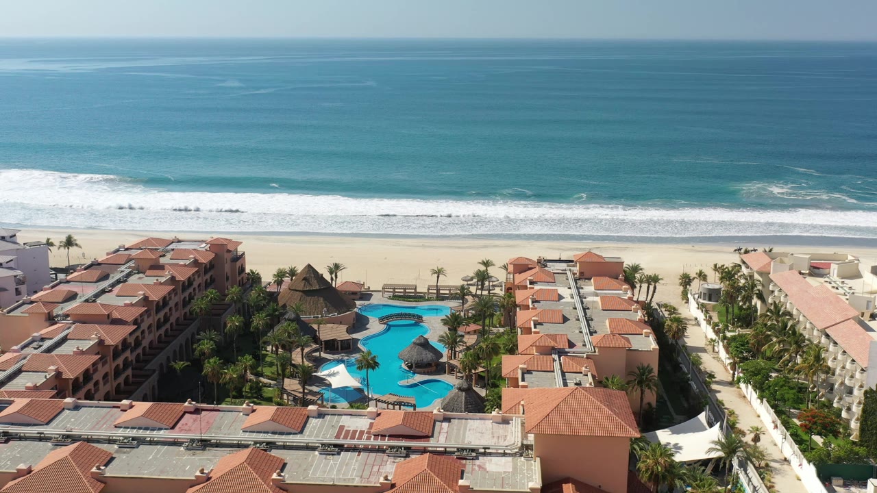 Aerial frame in Los Cabos, Mexico with the resort complex centered below, white surf rolling toward the sand, blue pools, palm trees, and no visible people.