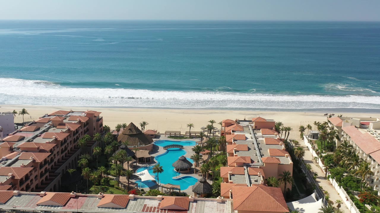 Aerial coastline with ocean, beach, resort rooftops, pools, and palms.