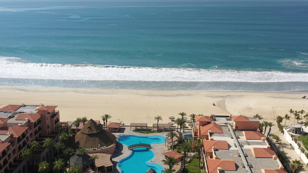 Aerial view of resort pools, orange roofs, sandy beach, and ocean waves.