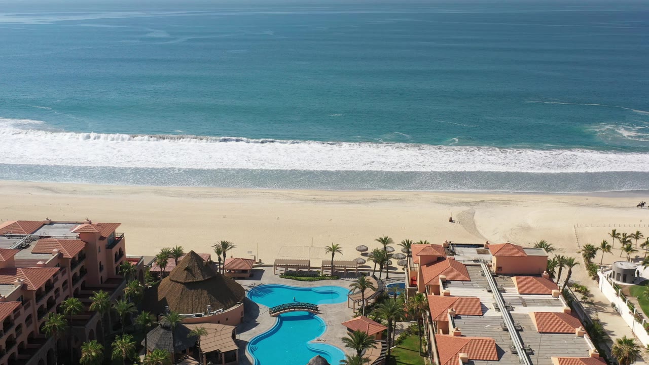 Elevated Los Cabos coastline view with the pool complex and orange tile roofs at the bottom, open sand through the center, and a long white wave line closer to shore.
