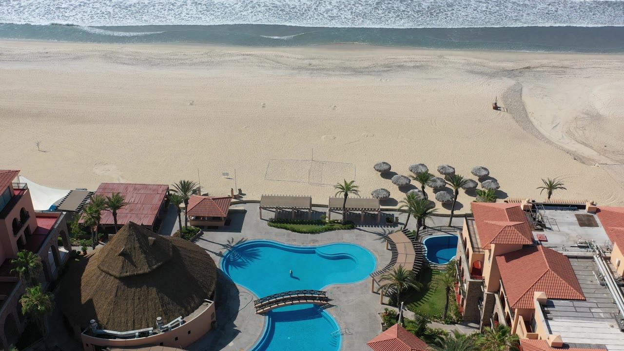 Aerial frame in Los Cabos, Mexico with a turquoise pool in the lower center, palapa-style umbrellas on the sand, volleyball court outlines, red roofs, palms, and no clearly recognizable people.