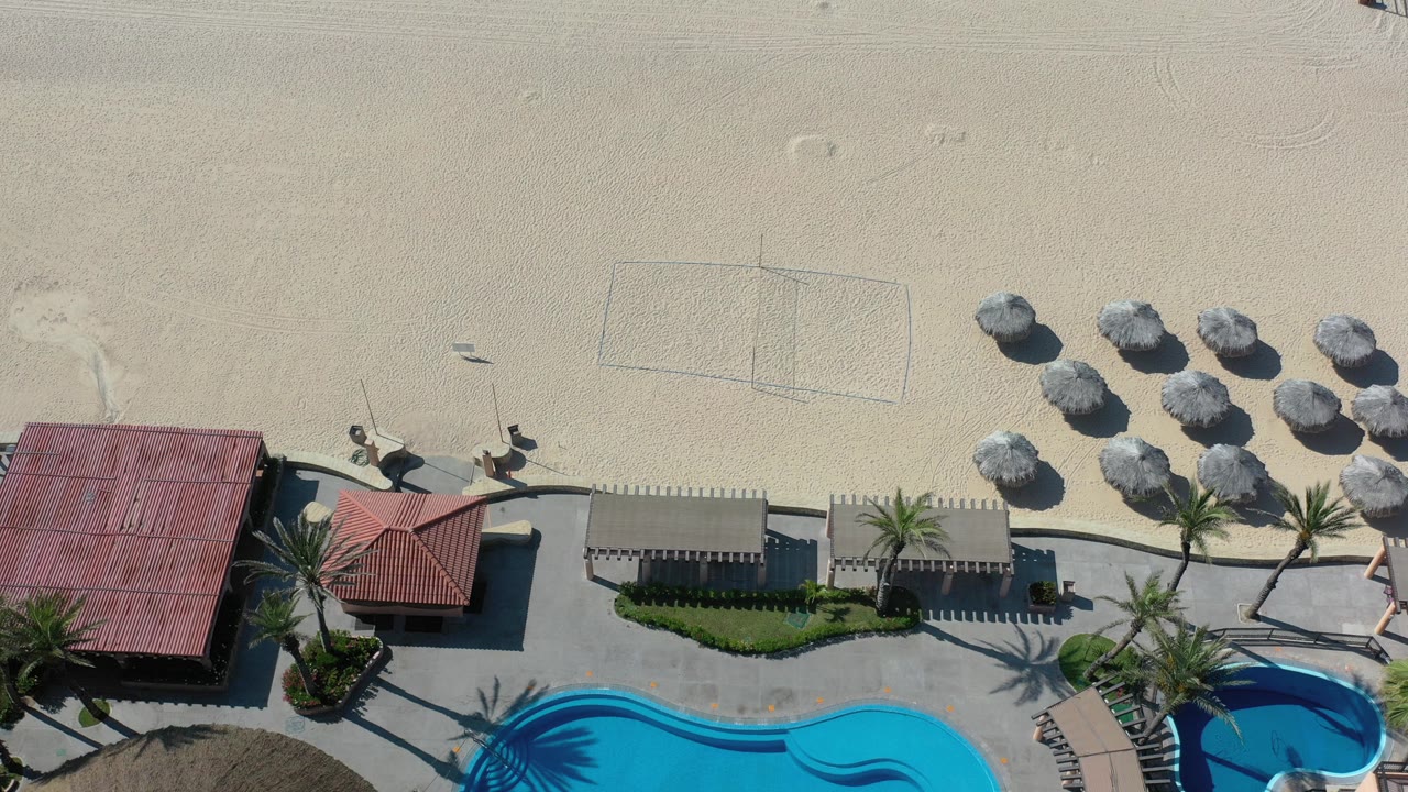 Overhead Los Cabos beach frame with red-roof beach structures, palm trees, a turquoise pool edge, shade umbrellas, a volleyball court on pale sand, and no visible people.