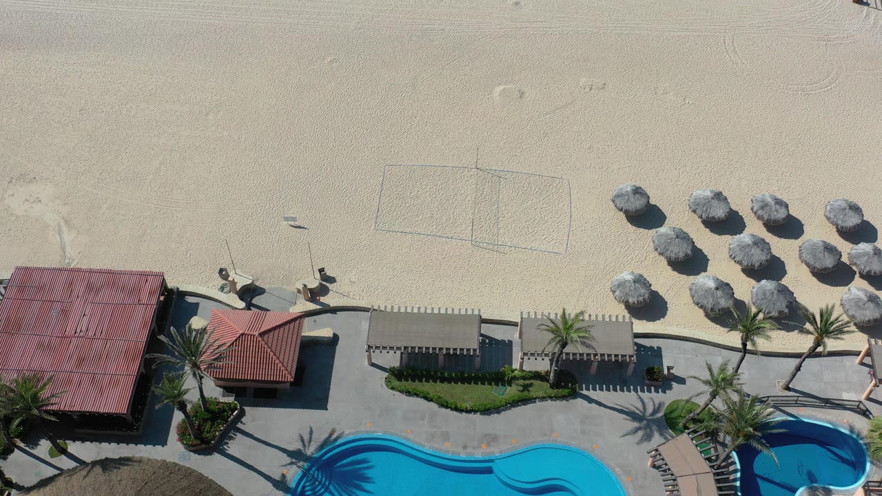 Los Cabos aerial view with red and tan beach roofs along the lower frame, palms casting shadows, a marked volleyball court in the sand, umbrellas, and no visible people.