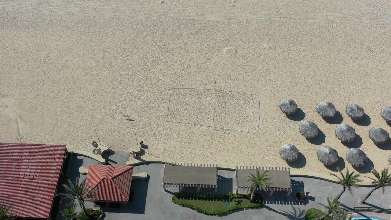 Top-down beach scene with volleyball court, umbrellas, palms, and red-roof structures.