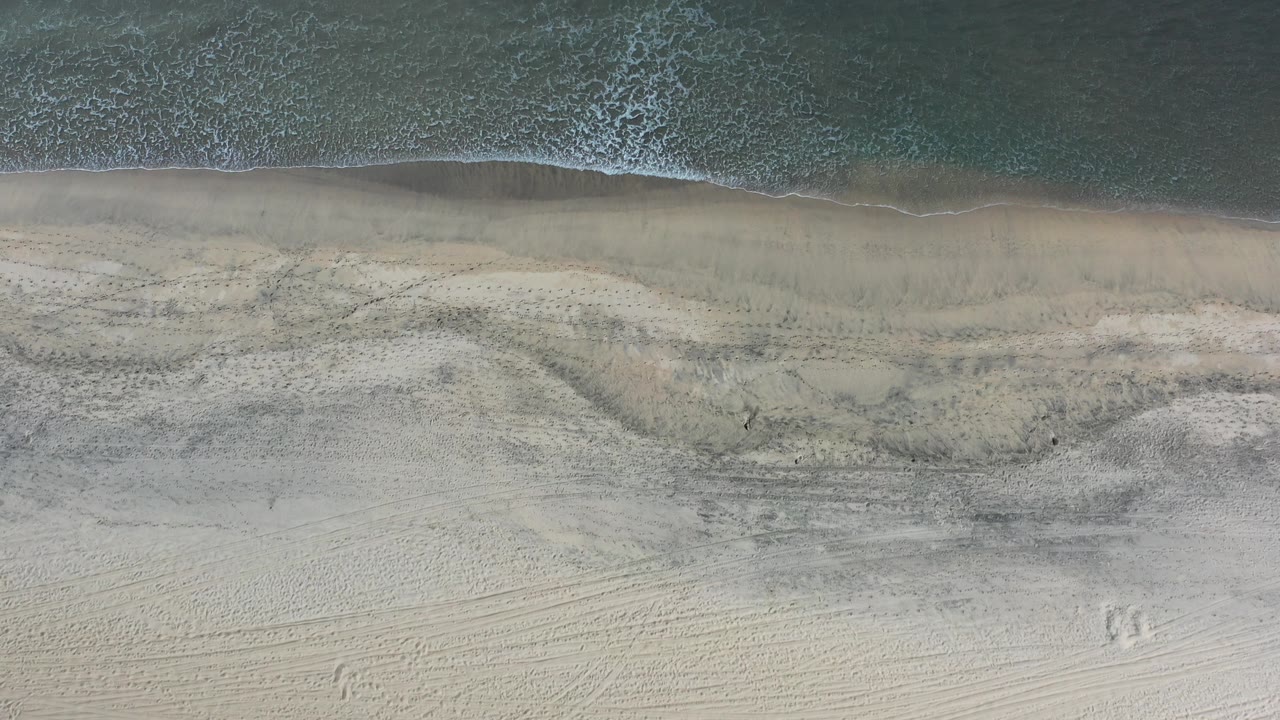 Overhead frame of a Los Cabos beach where a narrow wave line meets pale sand, with darker wet bands and no visible people.