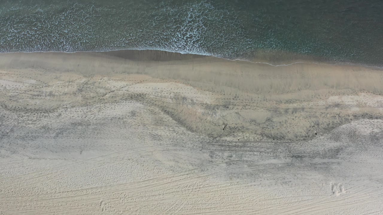 Top-down Los Cabos beach view with shallow green water along the upper edge, pale sand below, and no visible people.