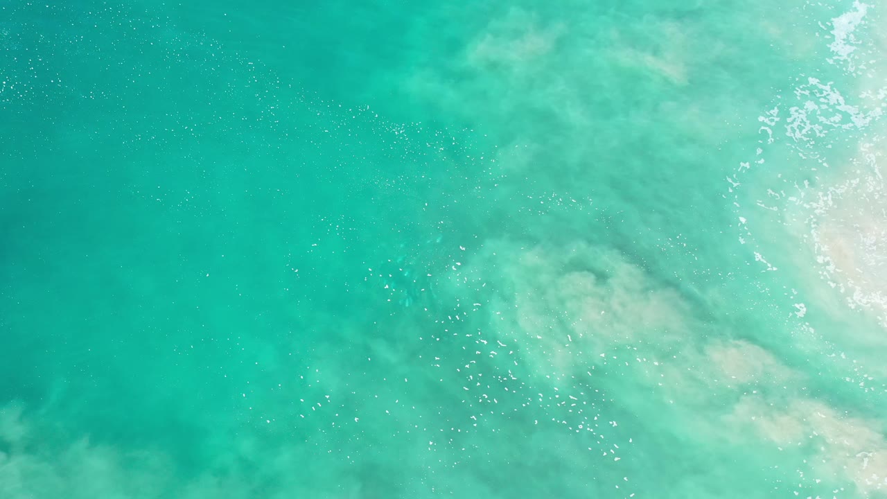 Overhead Los Cabos ocean frame with aqua water, a clouded sandy patch near center, fine bubbles across the shallows, bright foam along the right side, and no visible people.