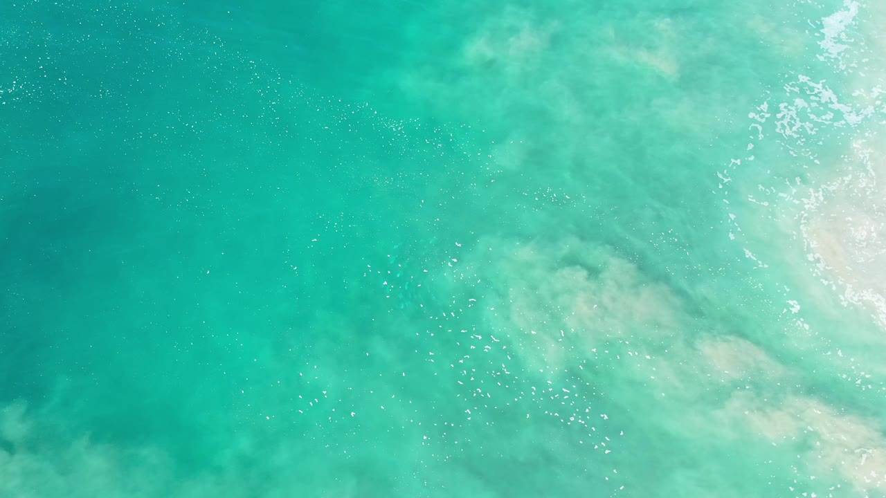 Clear teal water in Los Cabos fills the frame from above, with a pale sandy swirl under the surface, small bubble trails, broken white surf at right, and no visible people.
