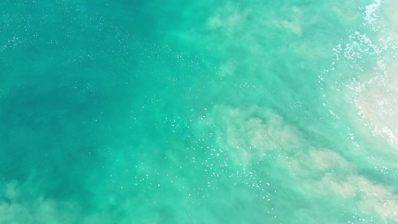 Overhead Los Cabos view of turquoise shallows where suspended pale sand softens the water color, white surf pushes in from the right edge, and no visible people appear.